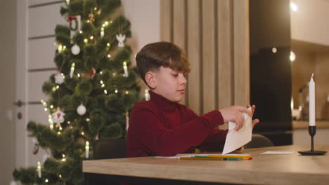 Junge-Im-Roten-Rollkragenpullover-Faltet-Einen-Wunschbrief-Und-Steckt-Ihn-In-Einen-Umschlag,-Der-An-Einem-Tisch-In-Einem-Mit-Einem-Weihnachtsbaum-Geschmückten-Raum-Sitzt