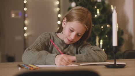 Chica-Con-Suéter-Verde-Escribiendo-Una-Carta-De-Deseos-Sentada-En-Una-Mesa-En-Una-Habitación-Decorada-Con-Un-árbol-De-Navidad