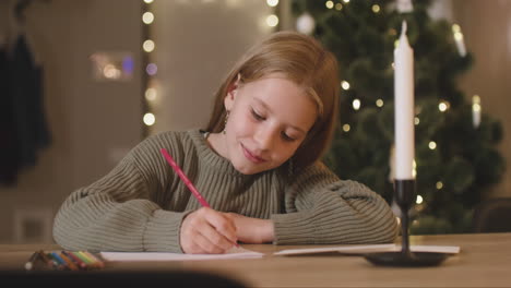 Chica-Con-Suéter-Verde-Escribiendo-Una-Carta-Y-Pensando-En-Deseos-Sentada-En-Una-Mesa-En-Una-Habitación-Decorada-Con-Un-árbol-De-Navidad