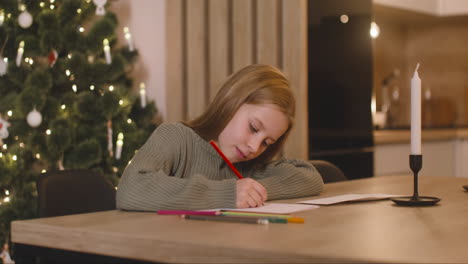 Chica-Con-Suéter-Verde-Escribiendo-Una-Carta-Y-Pensando-En-Deseos-Sentada-En-Una-Mesa-En-Una-Habitación-Decorada-Con-Un-árbol-De-Navidad-2