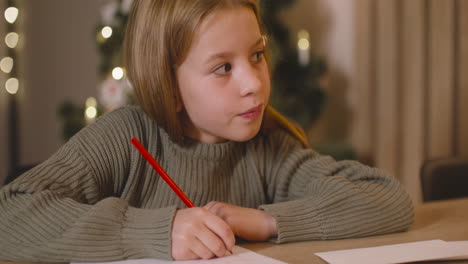 Close-Up-View-Of-A-Girl-In-Green-Sweater-Writing-A-Letter-And-Thinking-Of-Wishes-Sitting-At-A-Table-In-A-Room-Decorated-With-A-Christmas-Tree