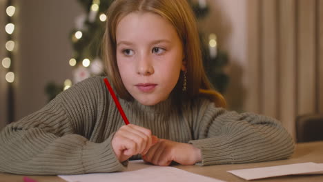 Nahaufnahme-Eines-Mädchens-Im-Grünen-Pullover,-Das-Einen-Brief-Schreibt-Und-An-Wünsche-Denkt,-Die-An-Einem-Tisch-In-Einem-Mit-Einem-Weihnachtsbaum-Geschmückten-Raum-Sitzen-1