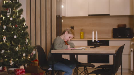 Side-View-Of-A-Girl-In-Green-Sweater-Writing-A-Letter-And-Thinking-Of-Wishes-Sitting-At-A-Table-In-A-Room-Decorated-With-A-Christmas-Tree