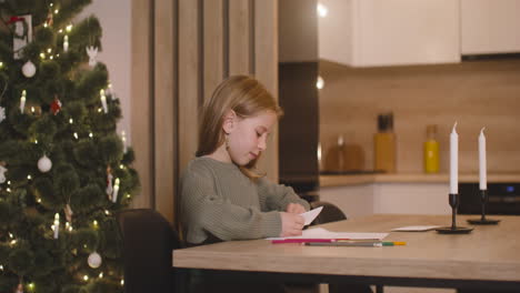 Girl-In-Green-Sweater-Folding-A-Letter-And-Inserting-It-In-A-Envelope-Sitting-At-A-Table-In-A-Room-Decorated-With-A-Christmas-Tree,-Then-Gets-Up-From-The-Chair-And-Leaves-The-Room