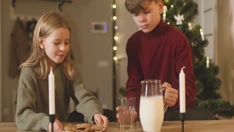 A-Blond-Boy-And-His-Blond-Sister-Place-An-Empty-Glass,-A-Jug-Of-Milk-And-A-Plate-Full-Of-Cookies-On-An-Empty-Table-With-Two-Candles,-Then-They-Leave-The-Room-1