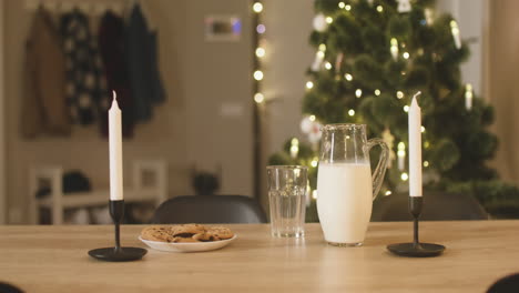 Camera-Focuses-On-An-Empty-Glass,-A-Jug-Of-Milk-And-A-Plate-Full-Of-Cookies-On-An-Empty-Table-With-Two-Candles-In-A-Room-Decorated-With-A-Christmas-Tree