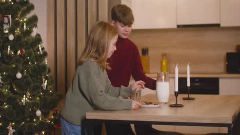 A-Blond-Boy-And-His-Blond-Sister-Place-An-Empty-Glass,-A-Jug-Of-Milk-And-A-Plate-Full-Of-Cookies-On-An-Empty-Table-With-Two-Candles,-Then-They-Leave-The-Room-2