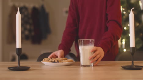 Camera-Focuses-On-A-Children's-Hands-Placing-A-Milk-Glass-And-A-Plate-Full-Of-Cookies-On-An-Empty-Table-With-Two-Candles-In-A-Room-Decorated-With-A-Christmas-Tree
