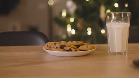 Camera-Focuses-On-An-Glass-Of-Milk-And-A-Plate-Full-Of-Cookies-On-An-Empty-Table-In-A-Room-Decorated-With-A-Christmas-Tree