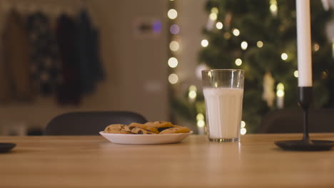Die-Kamera-Konzentriert-Sich-Auf-Ein-Glas-Milch-Und-Einen-Teller-Voller-Kekse-Auf-Einem-Leeren-Tisch-Mit-Zwei-Kerzen-In-Einem-Mit-Einem-Weihnachtsbaum-Geschmückten-Raum