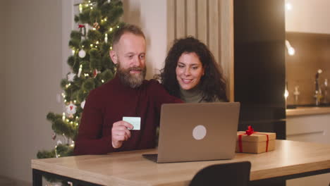 Ein-Paar-Kauft-Online-Mit-Einer-Kreditkarte-Mit-Einem-Laptop,-Das-An-Einem-Tisch-In-Der-Nähe-Eines-Geschenks-In-Einem-Mit-Einem-Weihnachtsbaum-Geschmückten-Raum-Sitzt