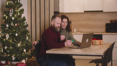 Seitenansicht-Eines-Paares,-Das-Mit-Einer-Kreditkarte-Online-Kauft-Und-Einen-Laptop-Verwendet,-Der-An-Einem-Tisch-In-Der-Nähe-Eines-Geschenks-In-Einem-Mit-Einem-Weihnachtsbaum-Geschmückten-Raum-Sitzt