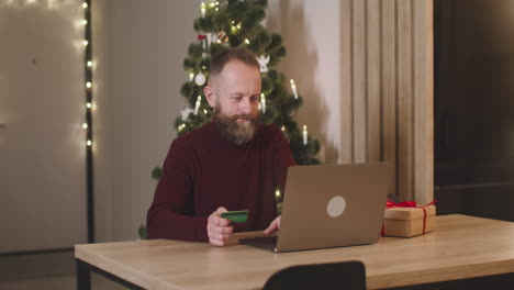 Rothaariger-Mann,-Der-Online-Mit-Einer-Kreditkarte-über-Einen-Laptop-Kauft,-Sitzt-An-Einem-Tisch-In-Der-Nähe-Eines-Geschenks-In-Einem-Mit-Einem-Weihnachtsbaum-Geschmückten-Raum-1