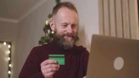 Unteransicht-Eines-Rothaarigen-Mannes,-Der-Online-Mit-Einer-Kreditkarte-über-Einen-Laptop-An-Einem-Tisch-In-Einem-Mit-Einem-Weihnachtsbaum-Geschmückten-Raum-Sitzt-1