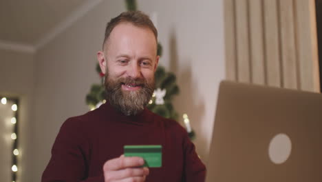 Unteransicht-Eines-Rothaarigen-Mannes,-Der-Online-Mit-Einer-Kreditkarte-über-Einen-Laptop-Kauft,-Der-An-Einem-Tisch-In-Einem-Mit-Einem-Weihnachtsbaum-Geschmückten-Raum-Sitzt-2