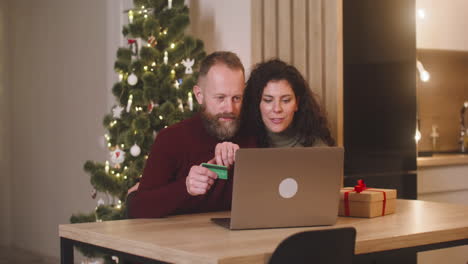 Paar,-Das-Online-Mit-Einer-Kreditkarte-über-Einen-Laptop-Kauft,-Sitzt-An-Einem-Tisch-In-Der-Nähe-Eines-Geschenks-In-Einem-Mit-Einem-Weihnachtsbaum-Geschmückten-Raum-1