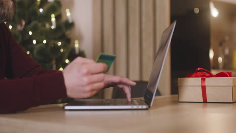 Side-View-Of-A-Man's-Hands-Using-Laptop-And-Holding-Credit-Card-Sitting-At-A-Table-Near-A-Present-In-A-Room-Decorated-With-A-Christmas-Tree