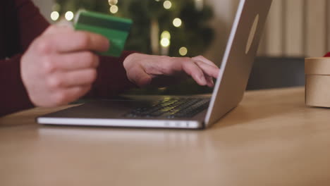 Camera-Focuses-On-Man's-Hands-Using-Laptop-And-Holding-Credit-Card-Sitting-At-A-Table-Near-A-Present-In-A-Room-Decorated-With-A-Christmas-Tree