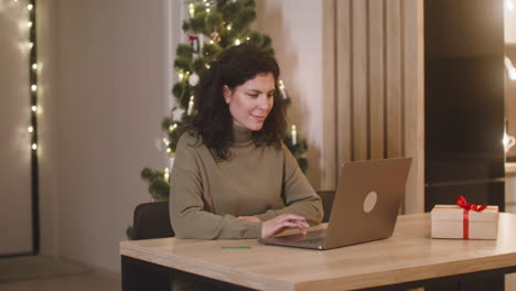 Brünette-Frau,-Die-Online-Mit-Einer-Kreditkarte-Mit-Einem-Laptop-An-Einem-Tisch-Neben-Einem-Geschenk-In-Einem-Mit-Einem-Weihnachtsbaum-Geschmückten-Raum-Kauft