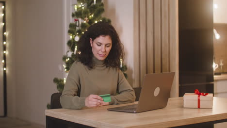 Brünette-Frau,-Die-Online-Mit-Einer-Kreditkarte-über-Einen-Laptop-An-Einem-Tisch-Neben-Einem-Geschenk-In-Einem-Mit-Einem-Weihnachtsbaum-Geschmückten-Raum-Kauft-1