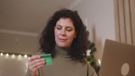 Bottom-View-Of-Brunette-Woman-Buying-Online-With-A-Credit-Card-Using-A-Laptop-Sitting-At-A-Table-In-A-Room-Decorated-With-A-Christmas-Tree