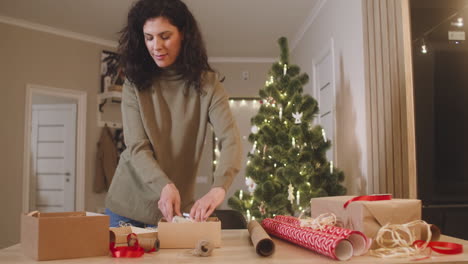 Brünette-Frau,-Die-Weihnachtsgeschenke-Auf-Einem-Tisch-In-Einem-Mit-Einem-Weihnachtsbaum-Geschmückten-Raum-Einwickelt