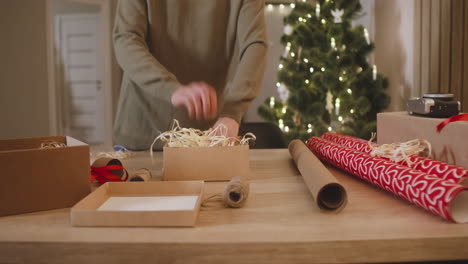 Die-Kamera-Konzentriert-Sich-Auf-Die-Hand-Der-Frau,-Die-Weihnachtsgeschenke-Auf-Einem-Tisch-In-Einem-Mit-Einem-Weihnachtsbaum-Geschmückten-Raum-Einwickelt