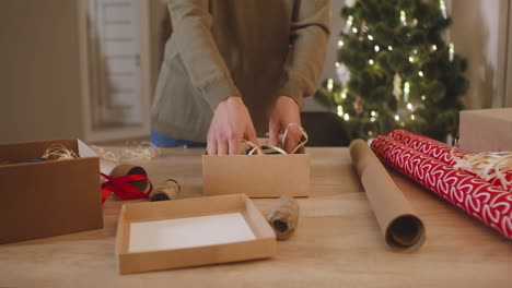 Die-Kamera-Konzentriert-Sich-Auf-Die-Hand-Einer-Frau,-Die-Weihnachtsgeschenke-Auf-Einem-Tisch-In-Einem-Mit-Einem-Weihnachtsbaum-Geschmückten-Raum-Einwickelt-1