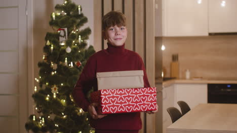 Vista-Frontal-De-Un-Niño-Rubio-Con-Suéter-De-Cuello-Alto-Rojo-Sosteniendo-Regalos-En-Una-Habitación-Decorada-Con-Un-árbol-De-Navidad