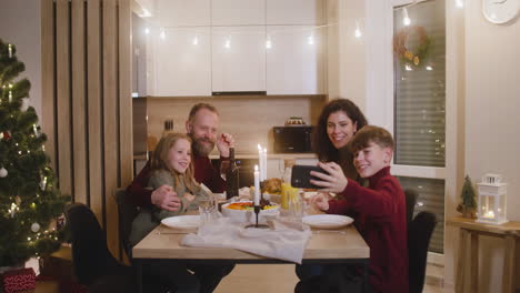 Der-Blonde-Junge,-Der-Mit-Seiner-Schwester-Und-Seinen-Eltern-Am-Weihnachtstisch-Sitzt,-Verwendet-Ein-Smartphone,-Um-Ein-Familien-Selfie-Zu-Machen