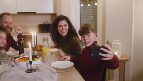 Blond-Boy-Sitting-With-His-Sister-And-Parents-At-The-Christmas-Dinner-Table-Uses-A-Smartphone-To-Make-A-Family-Selfie-2