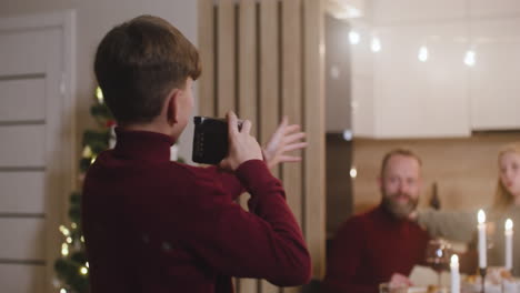 Ein-Blonder-Junge-In-Einem-Roten-Rollkragenpullover-Fotografiert-Seine-Eltern-Und-Seine-Schwester,-Die-Beim-Weihnachtsessen-Am-Tisch-Sitzen