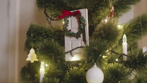 Camera-Focuses-On-White-Door-Decorated-With-Christmas-Wreath-Hanging-On-Christmas-Tree-With-Lights