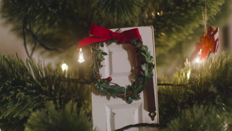 Camera-Focuses-On-White-Door-Decorated-With-Christmas-Wreath-Hanging-On-Christmas-Tree-With-Lights-1