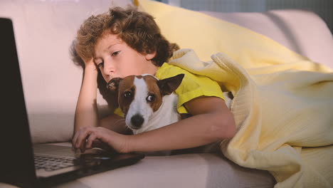 Blond-Boy-With-Curly-Hair-Using-Computer-While-Lying-With-His-Dog-On-The-Sofa