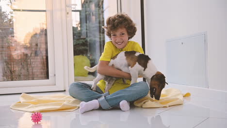 Blond-Boy-With-Curly-Hair-Sitting-On-The-Floor-Hugging-His-Dog