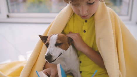 Blond-Boy-With-Curly-Hair-Sitting-On-The-Floor-Covered-With-A-Blanket-Next-To-His-Dog-While-Reading-1