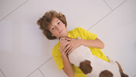 Top-View-Of-A-Blond-Boy-With-Curly-Hair-Lying-On-The-Floor-And-Caresses-His-Dog