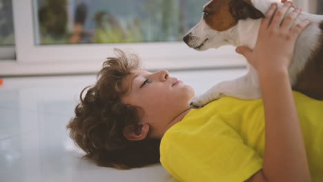 Side-View-Of-A-Blond-Boy-With-Curly-Hair-Lying-On-The-Floor-And-Caresses-His-Dog