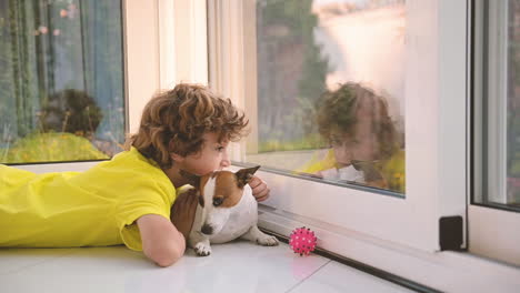 Blond-Boy-With-Curly-Hairs-Lying-On-The-Floor-With-His-Dog-While-Caresses-Him-And-Looking-Out-The-Window
