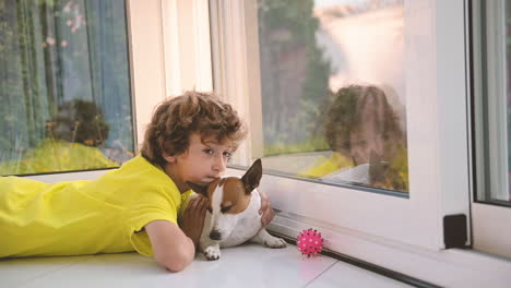 Blond-Boy-With-Curly-Hairs-Lying-On-The-Floor-With-His-Dog-While-Caresses-Him-And-Looking-Out-The-Window-1