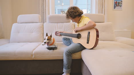 Blond-Boy-With-Curly-Hair-Playing-The-Guitar-Sitting-On-The-Couch,-Next-To-Him-Is-His-Dog-Lying