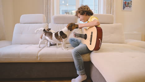 Blond-Boy-With-Curly-Hair-Playing-The-Guitar-Sitting-On-The-Couch,-Next-To-Him-Are-Their-Dogs-Lying-And-Walking-On-The-Sofa