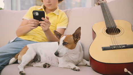 Blond-Boy-Sitting-On-The-Sofa-Using-A-Smartphone,-Next-To-His-Dog-Lying-Near-A-Guitar