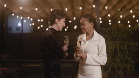 Two-Elegant-Multiethnic-Women-With-Champagne-Glasses-Talking-And-Laughing-At-New-Year's-Eve-Party