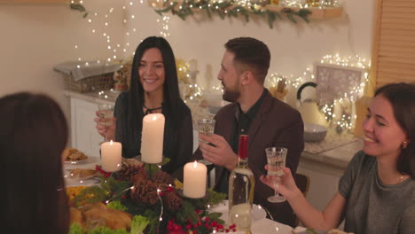 Group-Of-Happy-Friends-Sitting-At-Table,-Holding-Champagne-Glasses-And-Talking-To-Each-Other-At-Home-On-Christmas