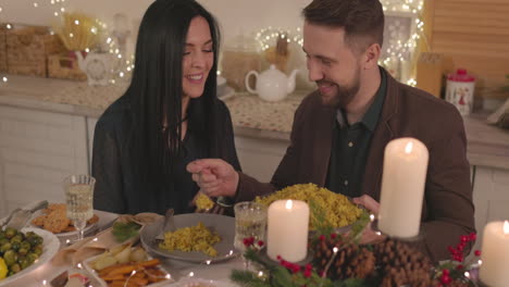 Smiling-Man-Sitting-At-Table-And-Putting-Rice-In-His-Girlfriend's-Plate-During-Christmas-Dinner
