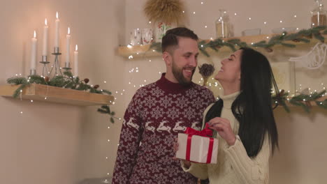 Happy-Woman-Holding-A-Christmas-Gift-Box-And-Kissing-Her-Boyfriend-At-Home