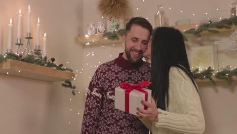 Happy-Woman-Holding-A-Christmas-Gift-Box-And-Kissing-Her-Boyfriend-At-Home-1