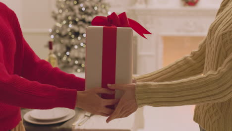 Unrecognizable-Woman-Giving-A-Gift-Box-To-Her-Friend-During-Christmas-Dinner-At-Home